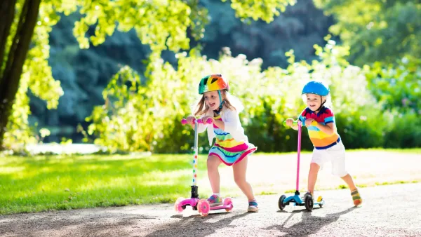 Two kids wearing helmets riding scoots on a summer day
